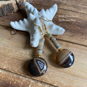 May include: A pair of agate earrings with gold-tone hooks and brown and white shell beads. The earrings are on a wooden surface with a white starfish.
