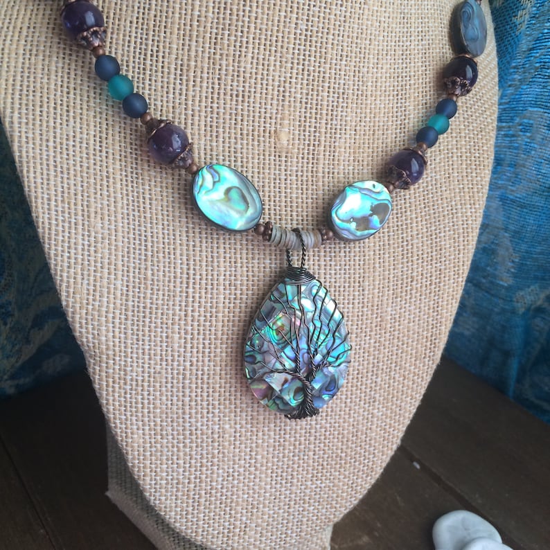 May include: A necklace with a large abalone shell pendant featuring a wire-wrapped tree design. The necklace is made with abalone shell beads, copper beads, and purple amethyst beads.