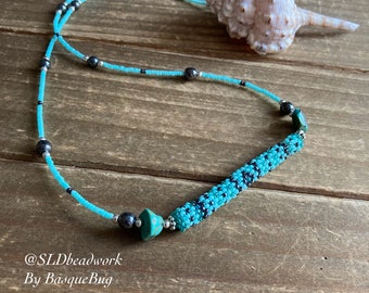 May include: A turquoise beaded necklace with a cylindrical central design, accented with black beads. The necklace features a delicate strand of turquoise beads and dark gray beads. The text "@SLDbeadwork By BasqueBug" is visible.