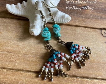 May include: Turquoise and brown beaded earrings with a tassel design. The earrings feature turquoise stones and brown beads, and the tassels are made of white, red, and turquoise beads. The earrings are hanging from a starfish on a wooden surface. The text "@SLDbeadwork By:BasqueBug" is visible in the image.