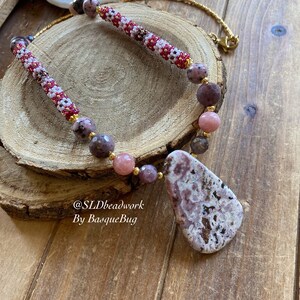 May include: A pink and purple gemstone necklace with a large pink and white agate pendant. The necklace is made with small beads and has a gold chain.