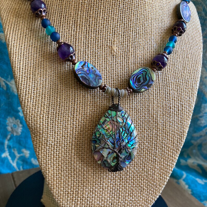 May include: A necklace with a large abalone shell pendant featuring a wire-wrapped tree design. The necklace is made with abalone shell beads, amethyst beads, copper beads, and blue glass beads.
