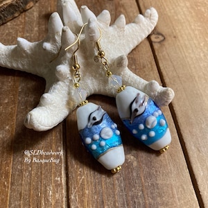 May include: A pair of dangle earrings with white oval glass beads. The beads have a blue and white design with a white dolphin on each bead. The earrings have gold hooks and a crystal bead on each hook.