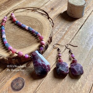 Può includere: Una collana di pietre preziose rosa, blu e viola con un paio di orecchini abbinati. La collana presenta un ciondolo di pietra preziosa grande e sfaccettato ed è realizzata con filo di rame e perline. Anche gli orecchini sono realizzati con filo di rame e perline e presentano pietre preziose sfaccettate più piccole.