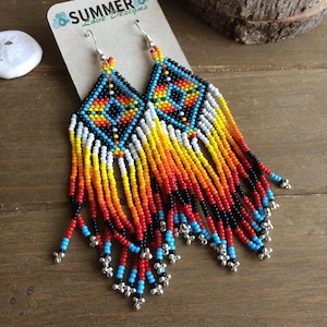 May include: A pair of long, beaded earrings with a geometric pattern in shades of black, white, red, orange, and blue. The earrings are hanging from silver hooks.
