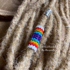 May include: A rainbow beaded dreadlock bead with white accents. The bead is on a brown dreadlock. The text "@SLDbeadwork By BasqueBug" is visible in the image.