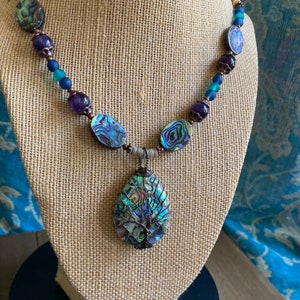 May include: A necklace featuring a large abalone shell pendant with a tree design. The necklace is made with abalone shell beads, purple glass beads, and copper wire.
