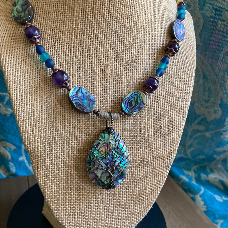 May include: A necklace featuring a large abalone shell pendant with a tree design. The necklace is made with abalone shell beads, purple glass beads, and copper wire.