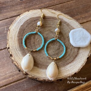 May include: A pair of turquoise and gold hoop earrings with a white seashell charm hanging from each hoop. The earrings are on a wooden surface with a white seashell in the background. @SLDbeadwork By BasqueBug