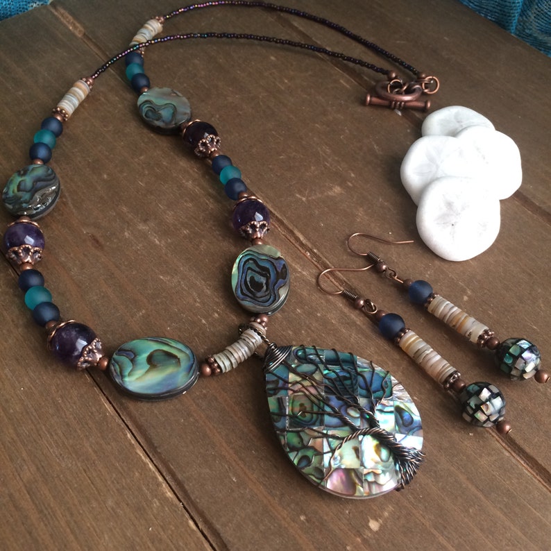 May include: A copper wire wrapped abalone shell pendant necklace and earrings set. The necklace features a teardrop shaped pendant with a tree design, abalone shell beads, amethyst beads, and copper beads. The earrings are copper wire wrapped abalone shell beads with copper beads.