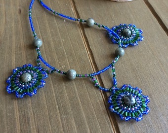 May include: A beaded necklace with three blue and green flower-shaped pendants, each with a gray stone in the center. The necklace is made with blue, green, and gray beads.