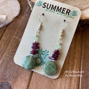 May include: A pair of dangle earrings with green and purple stone beads and a large green stone disc. The earrings are on a card that says "Summer Love Designs" and "@SLDbeadwork By BasqueBug".