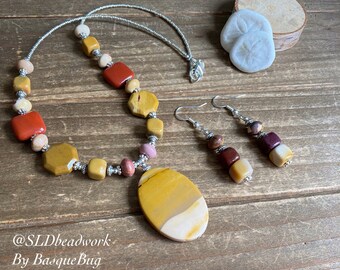 May include: A set of jewelry featuring a long necklace with a large oval pendant and matching earrings. The necklace and earrings are made with a variety of colorful stones, including red, yellow, and brown. The pendant is a large oval stone with a yellow, white, and brown pattern. The earrings are made with smaller stones in the same colors. The jewelry is on a wooden surface. @SLDbeadwork By BasqueBug
