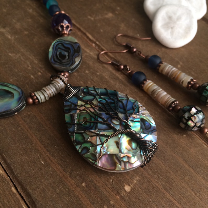 May include: A necklace and earrings set featuring a teardrop-shaped pendant with a wire-wrapped tree design. The pendant is made of iridescent abalone shell mosaic. The necklace and earrings are accented with copper beads and abalone shell beads.