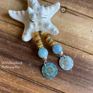 May include: A pair of dangle earrings with a blue and brown color scheme. The earrings feature a round blue stone, a stack of brown and white beads, and a silver charm with a blue and green mandala design.