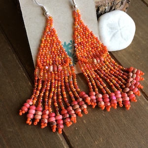 May include: A pair of handmade orange beaded earrings with silver hooks. The earrings feature a triangular design with multiple strands of orange beads, accented with pink and brown beads. The earrings are displayed on a wooden surface.