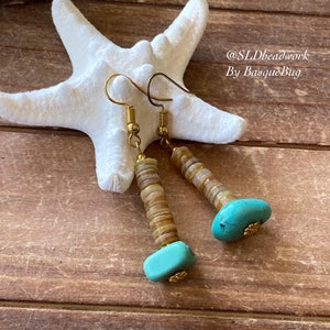 May include: A pair of gold tone earrings with turquoise stones and brown shell beads. The earrings are hanging from a white starfish on a wooden surface. @SLDbeadwork By BasqueBug