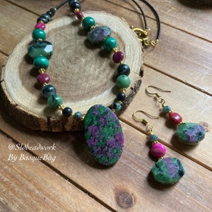 May include: A green and pink gemstone necklace and earrings set. The necklace features a large oval pendant and is made with round and faceted beads. The earrings are dangle style with a smaller oval gemstone.