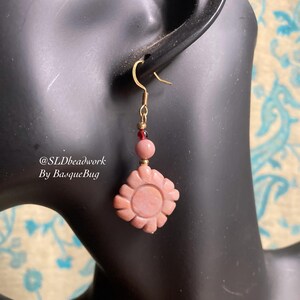 Op de afbeelding: Roze bloemvormige oorbellen met gouden accenten en rode kralen. @SLDbeadwork By BasqueBug