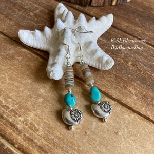 May include: Turquoise and brown shell bead earrings with silver nautilus shell charms. The earrings are hanging from a white starfish on a wooden surface. The text '@SLDbeadwork By:BasqueBug' is visible in the image.
