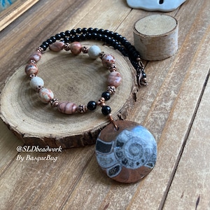 Może przedstawiać: Naszyjnik z miedzianym wisiorem przedstawiającym skamieniałość amonita. Naszyjnik wykonany jest z czarnych i brązowych koralików.