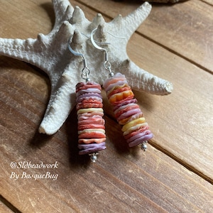 May include: A pair of dangle earrings made with stacked, colorful, shell-like beads. The beads are in shades of pink, orange, yellow, and purple. The earrings are silver-toned and have a hook closure.
