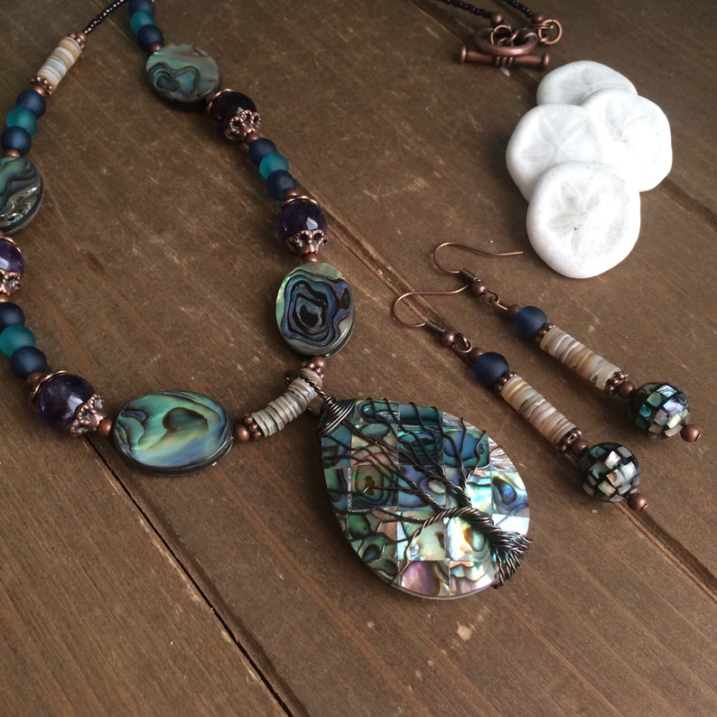 May include: A necklace and earrings set featuring a teardrop-shaped pendant with a wire-wrapped tree design. The pendant and earrings are made with iridescent abalone shell, copper wire, and blue and white glass beads.