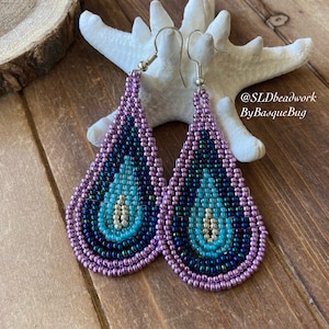 May include: A pair of teardrop-shaped beaded earrings with a multi-colored design. The earrings are made with purple, blue, turquoise, and silver beads. The earrings are hanging from silver hooks. The earrings are on a wooden surface with a white starfish.
