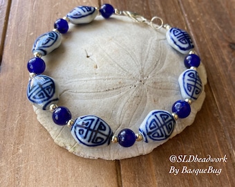 Pode incluir: Um bracelete de contas de porcelana azul e branco com um fecho prateado. As contas apresentam um design chinês tradicional. O bracelete está sobre um fundo branco.