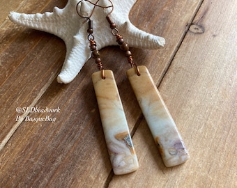 Peut inclure: Une paire de longues boucles d'oreilles rectangulaires en pierre naturelle et fil de cuivre. La pierre est de couleur brun clair, blanc et beige avec un motif marbré. Les boucles d'oreilles sont suspendues à un fil de cuivre avec de petites perles de cuivre. Les boucles d'oreilles sont sur une surface en bois avec une étoile de mer blanche.