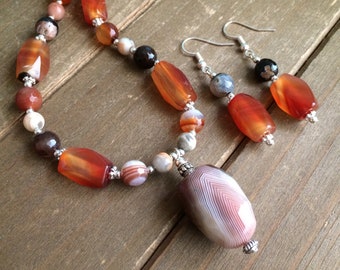 May include: A necklace and earring set featuring a variety of gemstone beads in shades of orange, brown, and white. The necklace has a large, striped pendant. The earrings have matching orange beads. The jewelry is displayed on a wooden surface.