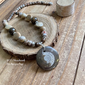 Może przedstawiać: Naszyjnik z zawieszką z amonita skamieniałego w kolorze brązowym i białym, srebrnym łańcuszkiem i brązowymi i białymi koralikami. Naszyjnik leży na drewnianej powierzchni. @SLDbeadwork By BasqueBug