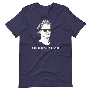 May include: A navy blue t-shirt featuring a white illustration of a Roman bust wearing sunglasses and a laurel wreath. The text "COOLIUS CAESAR" is printed below the bust.