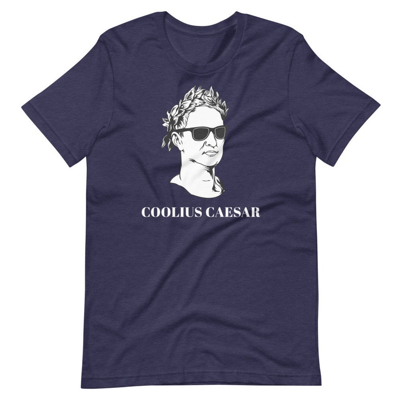 May include: A navy blue t-shirt featuring a white illustration of a Roman bust wearing sunglasses and a laurel wreath. The text "COOLIUS CAESAR" is printed below the bust.