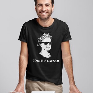 May include: A black t-shirt featuring a white graphic of a man wearing a laurel wreath and sunglasses. The text "COOLIUS CAESAR" is printed below the graphic.