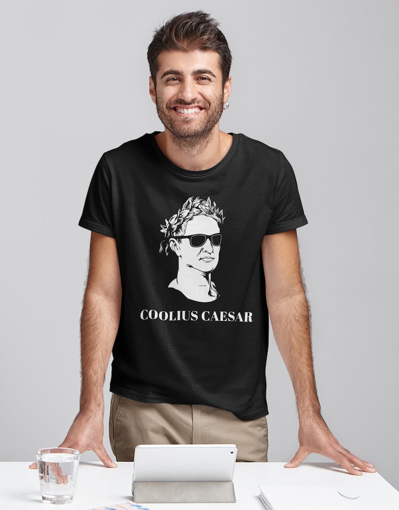May include: A black t-shirt featuring a white graphic of a man wearing a laurel wreath and sunglasses. The text "COOLIUS CAESAR" is printed below the graphic.