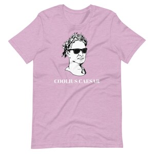 May include: A pink t-shirt featuring a black and white illustration of a bust of a man wearing sunglasses and a laurel wreath. The text "COOLIUS CAESAR" is printed below the illustration.