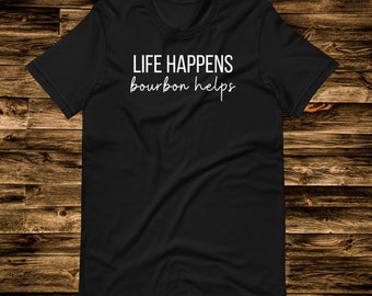 May include: Black t-shirt with the white text "LIFE HAPPENS bourbon helps" in a casual font. The shirt is displayed on a wooden surface. The t-shirt is a crew neck style.