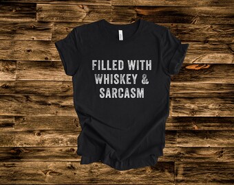 May include: Black t-shirt with the text "FILLED WITH WHISKEY & SARCASM" in distressed white lettering. The shirt is made of a soft material and is displayed on a wooden background.