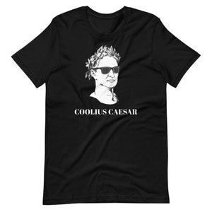 May include: Black t-shirt featuring a white illustration of a Roman bust wearing sunglasses and a laurel wreath. The text "COOLIUS CAESAR" is printed below the bust.