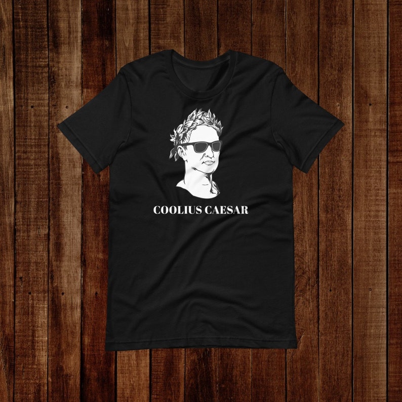 May include: Black t-shirt featuring a white graphic of a person wearing a laurel wreath and sunglasses. The text "COOLIUS CAESAR" is printed below the graphic.