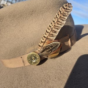 May include: A tan felt hat featuring a beige grosgrain ribbon band. The hat is decorated with a brass shotgun shell casing base and a cluster of brown and tan striped feathers tucked into the ribbon. The shell casing is marked with the text FEDERAL.
