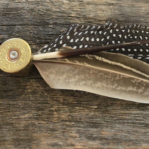Puede incluir: Broche artesanal con una vaina de cartucho de latón, adornada con una colección de plumas. Las plumas incluyen una pluma moteada en blanco y negro y una pluma de color marrón y crema. El broche está sobre un fondo de madera rústica.