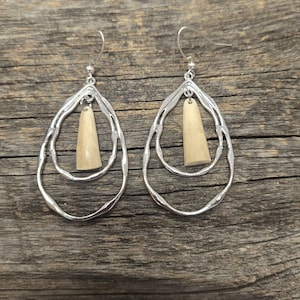 May include: A pair of silver-coloured teardrop earrings with a textured, wavy design. Each earring has a light beige, cone-shaped pendant suspended within the teardrop frame. The earrings are displayed on a weathered wooden surface.