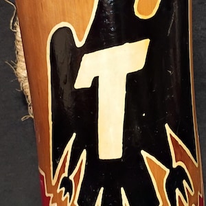 May include: A cylindrical wooden cup with a natural wood finish. The cup features a black eagle design with a white "T" in the center, outlined in cream. The bottom is painted red, and a natural fiber cord is attached.