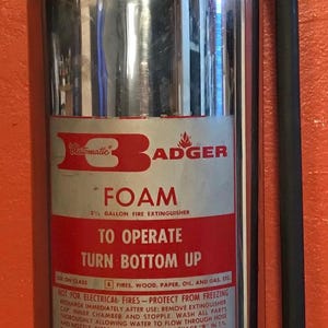 May include: A vintage Badger foam fire extinguisher, repurposed as a lamp. The silver-colored tank has a red top and a black hose. The label reads "FOAM" and "To Operate Turn Bottom Up". A light bulb is mounted on top.