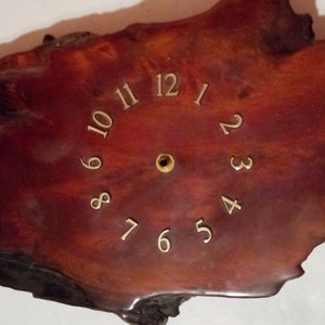 May include: A wooden clock face with gold numbers 1 through 12. The clock face is a natural wood shape with a reddish-brown color.