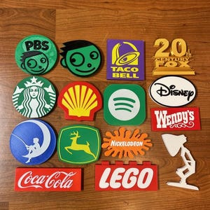 May include: A collection of colorful logo-shaped coasters, including PBS, Taco Bell, Disney, and Coca-Cola. The coasters are made of a solid material and are arranged on a wooden surface. The logos are in various shapes and colors.
