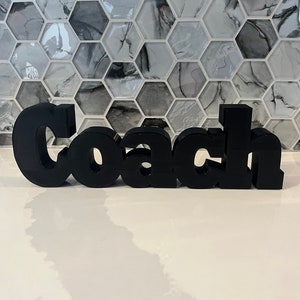 May include: Black 3D word "Coach" made of plastic. The word is sitting on a white surface.