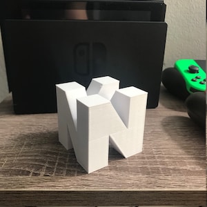May include: A white 3D printed model of the Nintendo logo, "N", on a wooden surface. The logo is made up of several interlocking cubes.